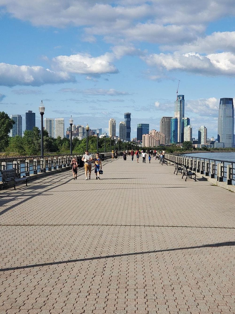 People walking along a riverside promenade with the New York City skyline in the background.