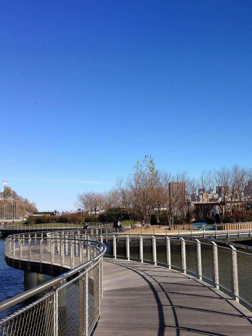 Scenic riverside park walkway with railing, trees, and city skyline under clear blue sky.
