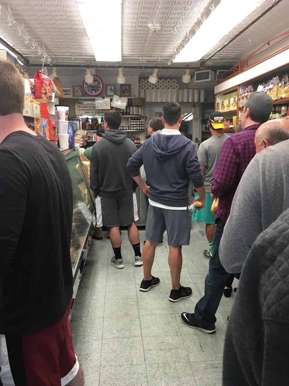 Crowd of people waiting in line at a bakery or deli, modern interior, casual clothing, busy food service environment.