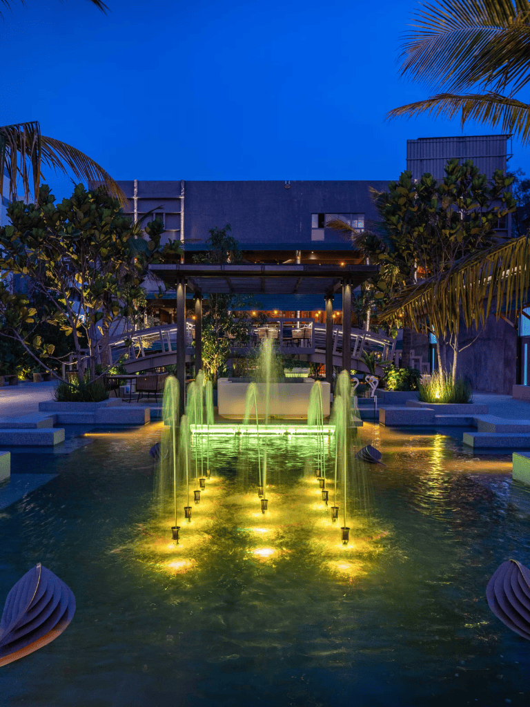 Elegant outdoor water fountain with colorful lighting at dusk, surrounded by lush tropical plants.
