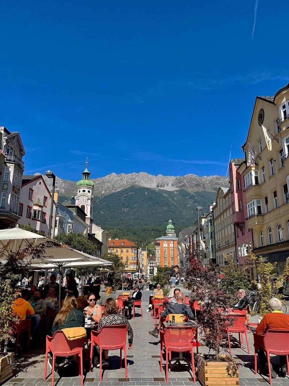 Colorful European city street with outdoor cafes, historic architecture, and mountain backdrop.