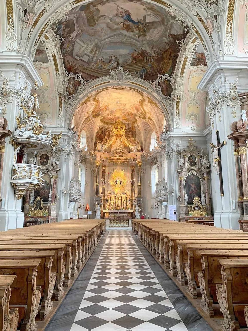 Ornate Baroque church interior with intricate white and gold decorations, painted ceiling, and wooden pews.