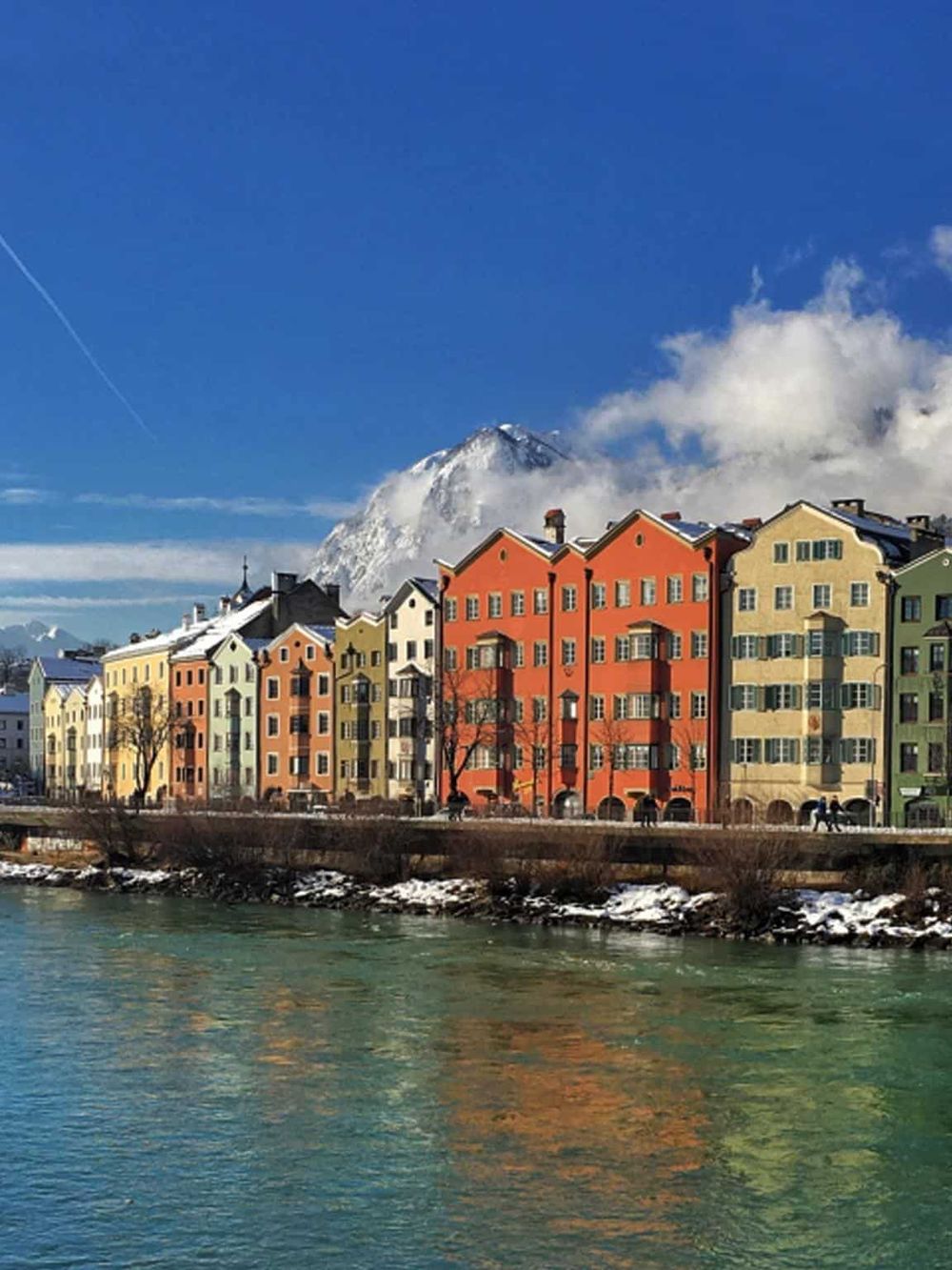 Colorful European-style buildings along a river with snowy mountain in background, scenic travel destination.