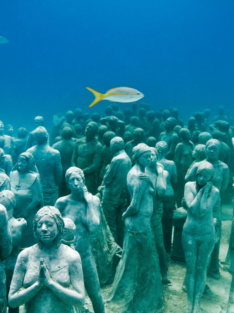People sculpture underwater in an art installation, with a fish swimming above the statues in the ocean.