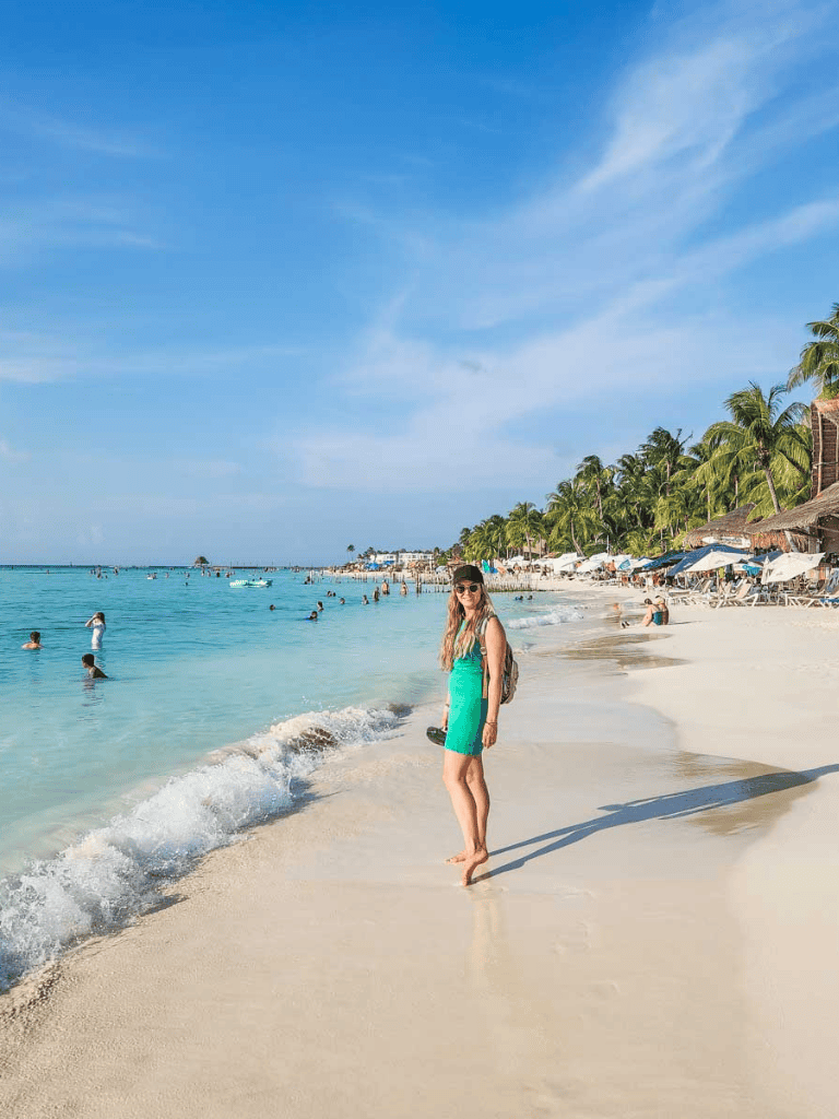 Vibrant beach scene with woman in turquoise dress, palm trees, and clear blue ocean on sunny day.