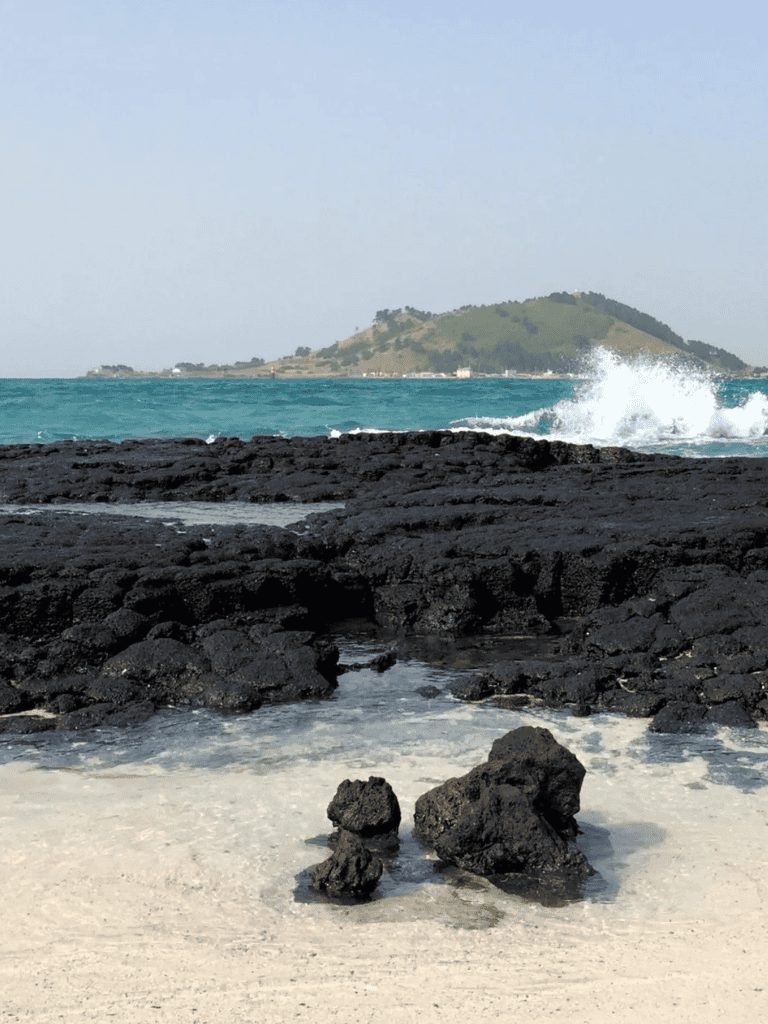 Sea rocks on beach with distant island and ocean waves, scenic coastal view - QuestForDirections.