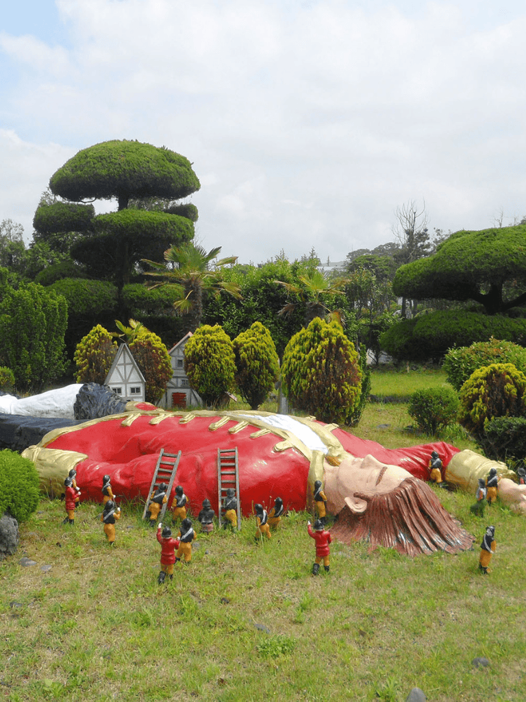 Brightly colored giant statue of a sleeping woman lying on the grass, surrounded by miniature figures and lush greenery.