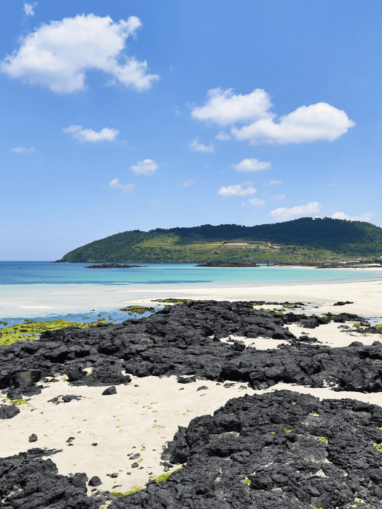 Vibrant beach scene with black rocks, white sands, and lush green hills under a bright blue sky.