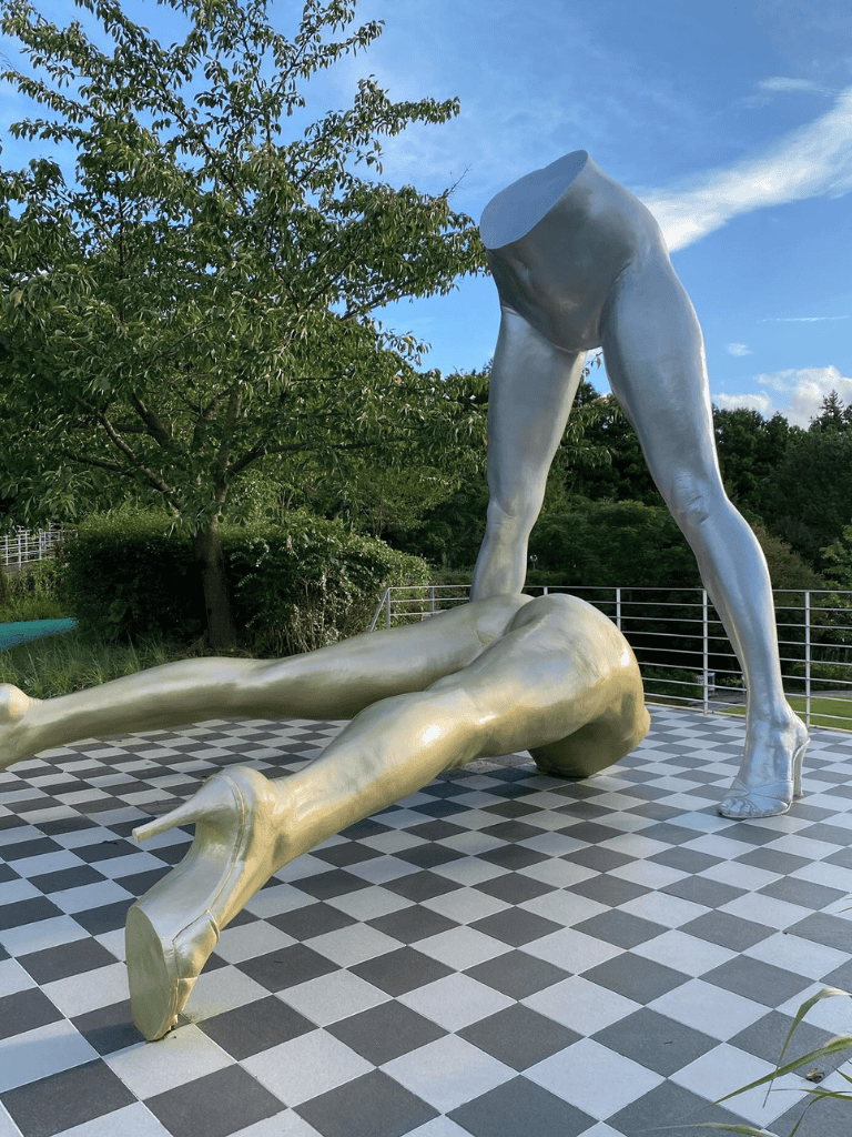 Metal sculpture of a headless woman with high heels lifting a fallen, nude woman on a checkered floor, outdoors under blue sky.