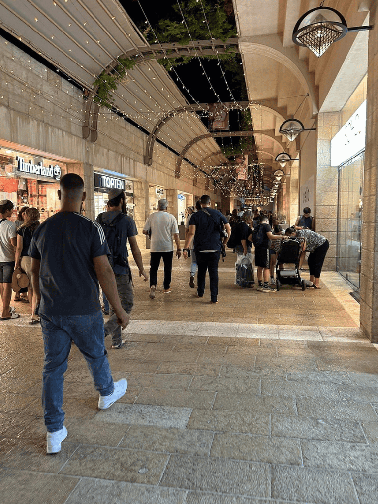 Busy shopping mall corridor with shoppers, stores, and ceiling string lights, captured for QuestForDirections.