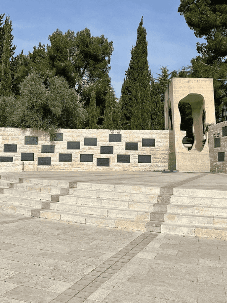 Empty outdoor stage with stone steps and modern sculpture, surrounded by tall trees, in a scenic park setting.