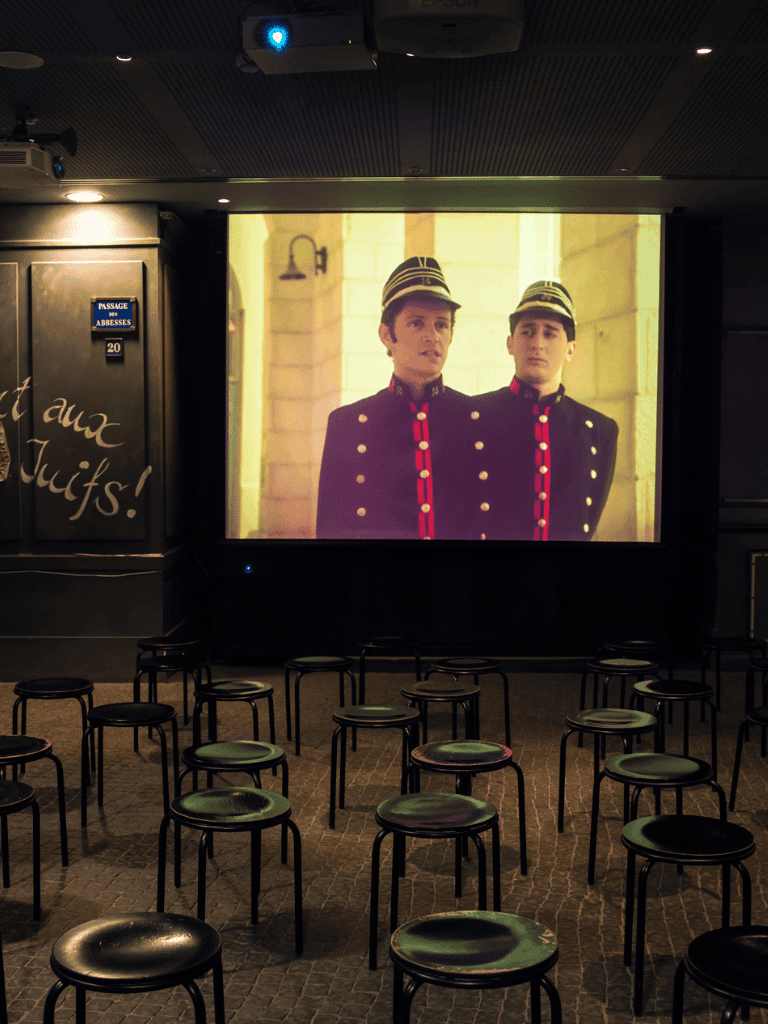 Loudspeakers and projection screen showing a man in hotel uniform with chairs in front of the screen in a lounge area.