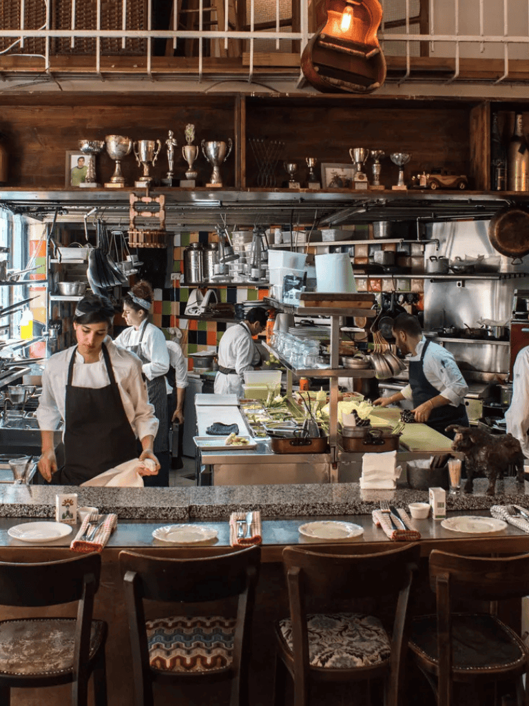 Busy restaurant kitchen with chefs preparing dishes, wooden shelves with trophies, and a cozy dining area in the foreground.