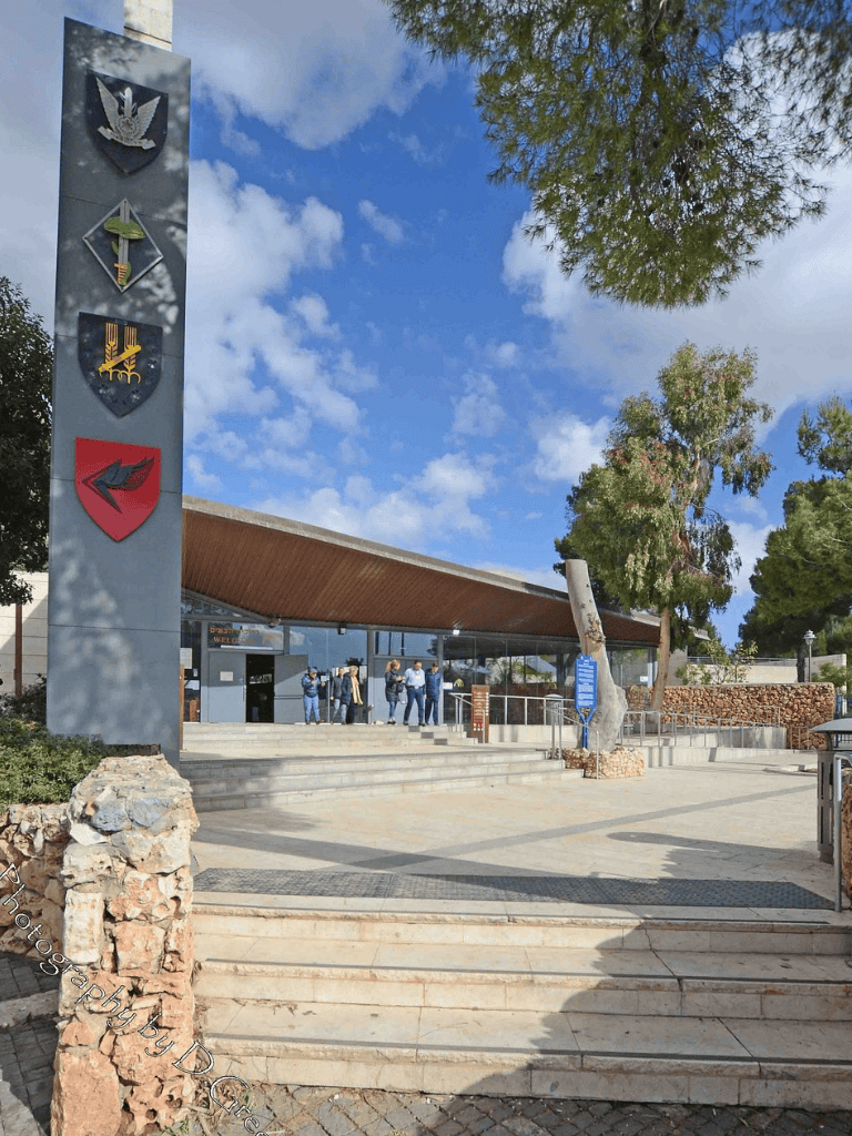 U.S. military entrance gateway featuring emblems on a tall concrete pillar, with visitors entering under a modern wooden canopy against a blue sky.