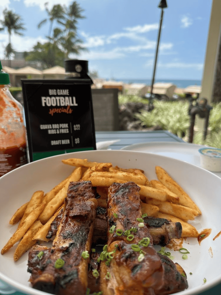 Grilled BBQ ribs with fries on a beachside patio, ocean view, palm trees, and sunny sky in the background.