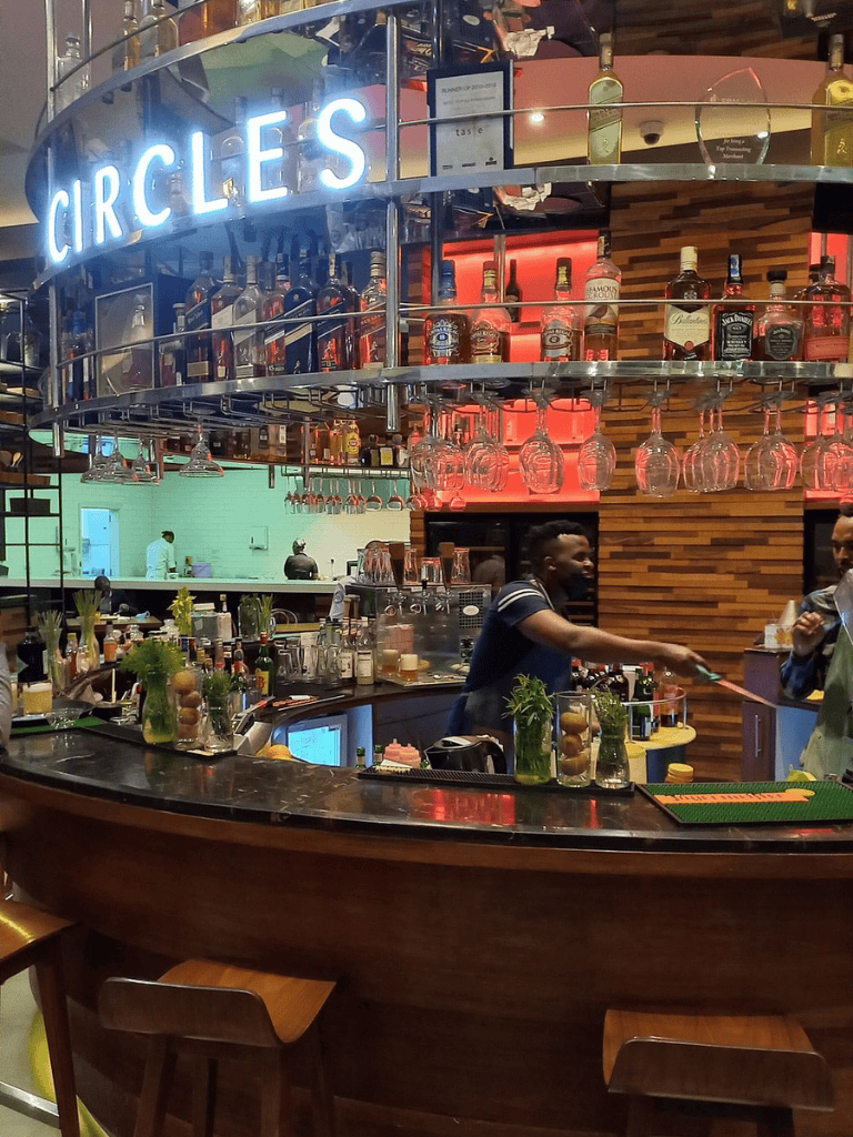Brightly lit bar at Circles restaurant with bartenders serving drinks, glassware hanging overhead, and neon signage.