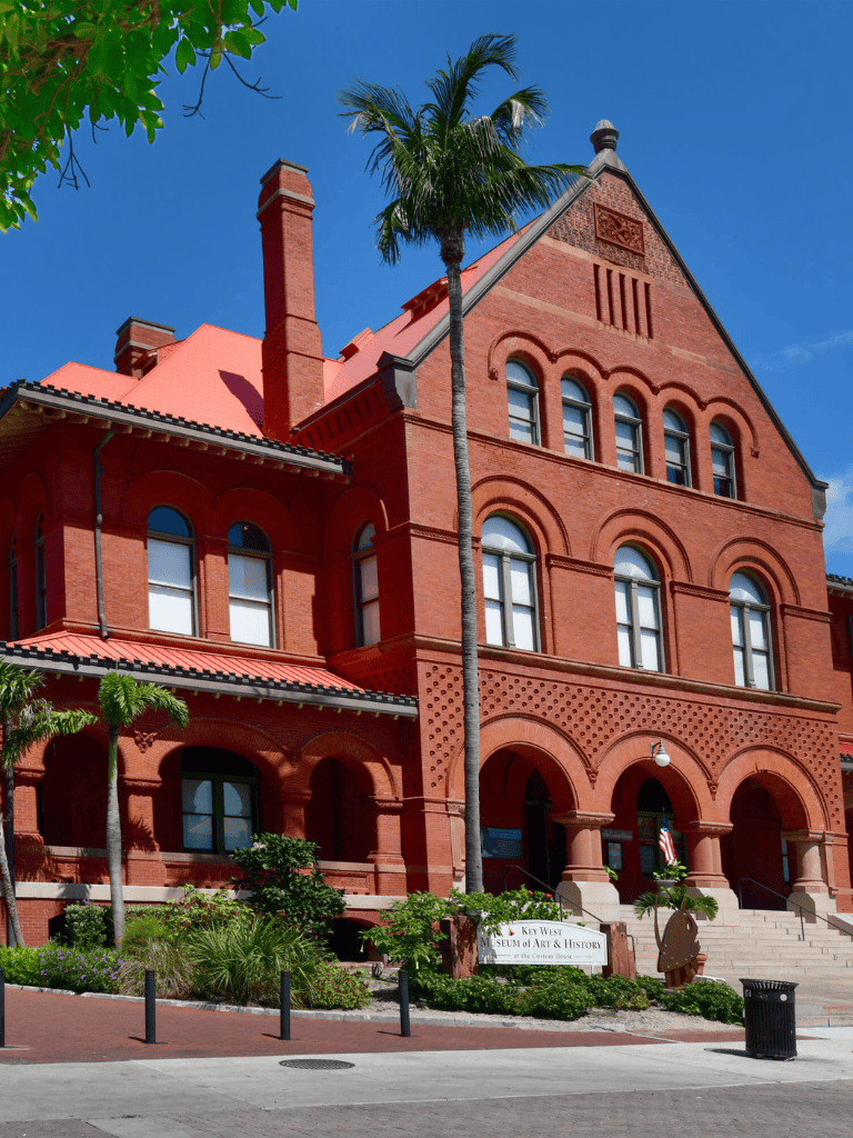 Bright red historic building with arched windows and palm trees, situated in Key West Museum of Art & History.