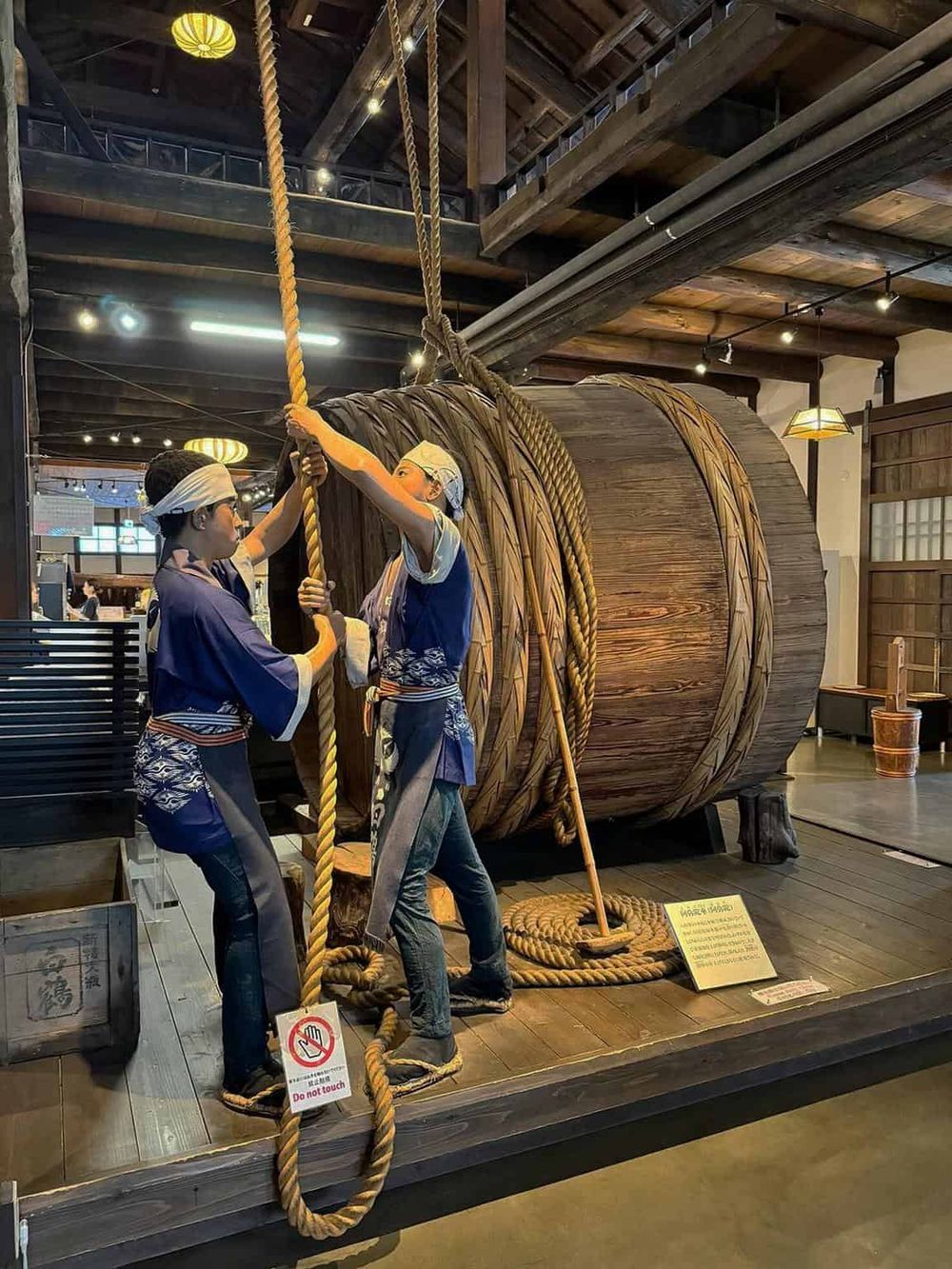 A giant wooden sake barrel with two life-sized figures of traditional brewers pulling a giant rope, showcasing Japanese sake brewing culture.