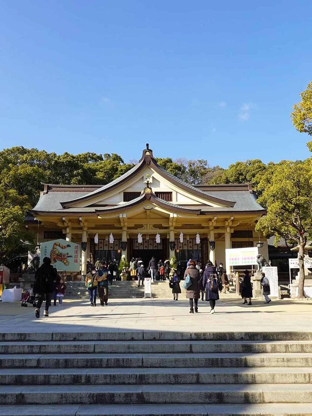 Serene Japanese shrine with visitors, surrounded by lush green trees on a sunny day.
