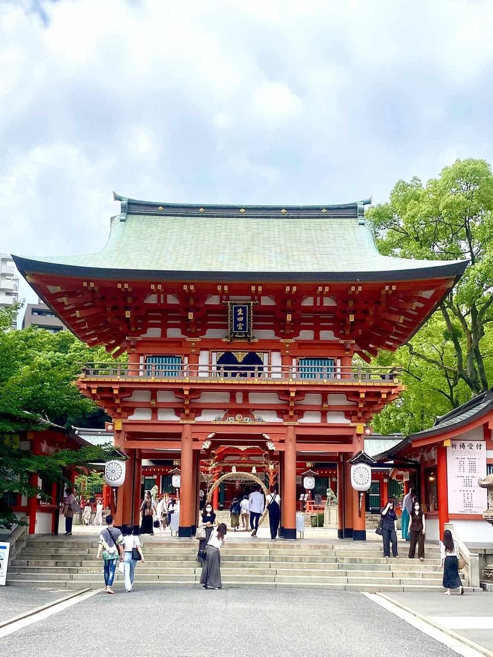Bright red traditional Japanese shrine with visitors, lush green trees, and modern buildings in background.