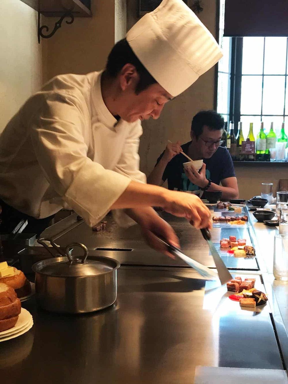 Experienced chef preparing sushi in a modern restaurant kitchen with a customer enjoying a bowl of ramen.