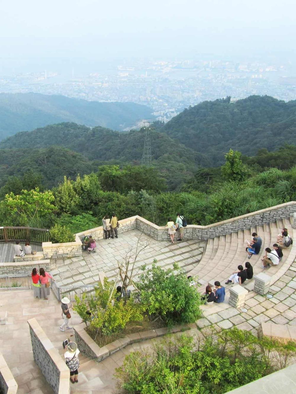 Panoramic view of mountain observation deck overlooking lush green hills and distant cityscape, popular outdoor sightseeing spot.
