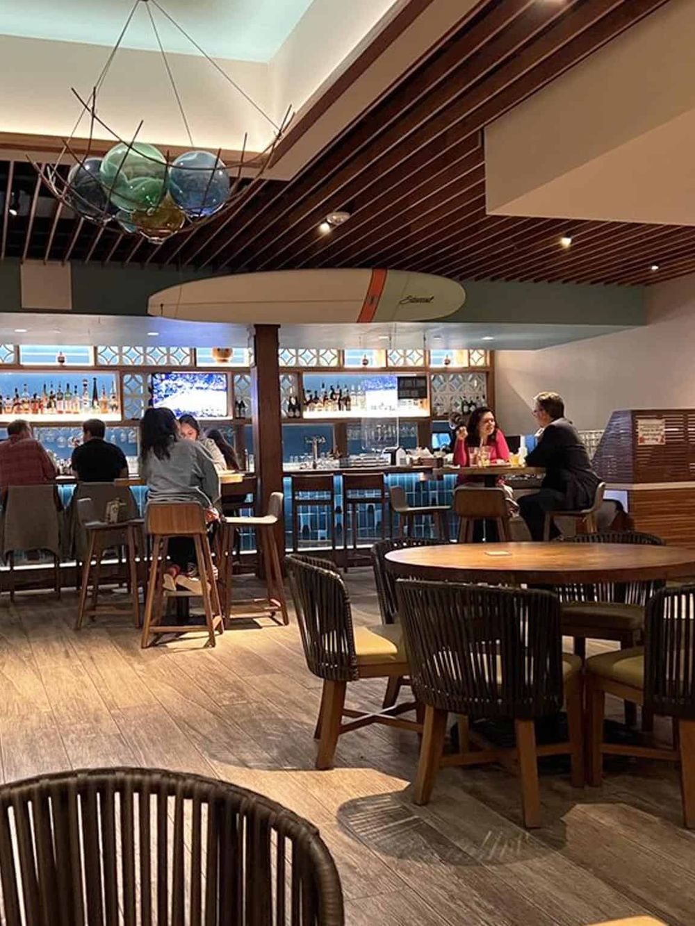 Modern restaurant interior with people dining, bar area, and surfboard decor, ideal for travel and food enthusiasts.