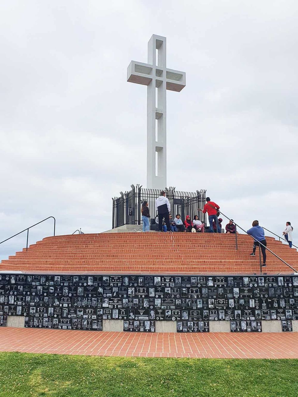 Large white cross monument at Quest for Directions site with visitors, memorial plaques, and cloudy sky.