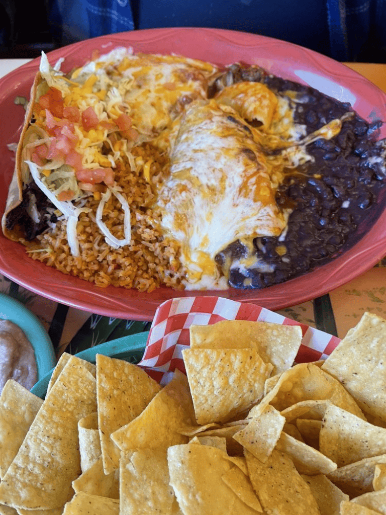 Shredded beef enchiladas with melted cheese, black beans, Mexican rice, and tortilla chips on a red plate.