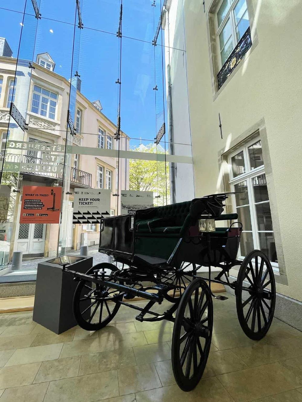 Vintage horse-drawn carriage display inside QuestForDirections museum with glass facade and historic city buildings background.
