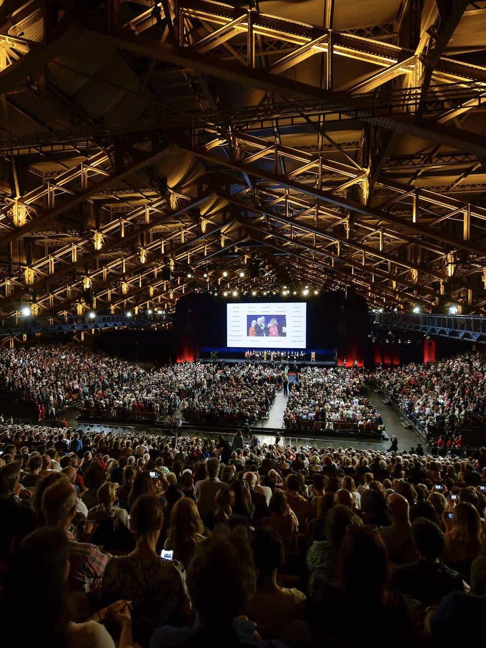 Brightly lit conference hall with a large audience and a stage featuring a presentation screen, emphasizing event planning and corporate gatherings.
