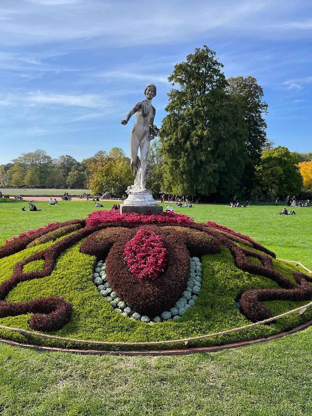 Statue of a woman in a park setting, surrounded by colorful flower bed and lush trees.