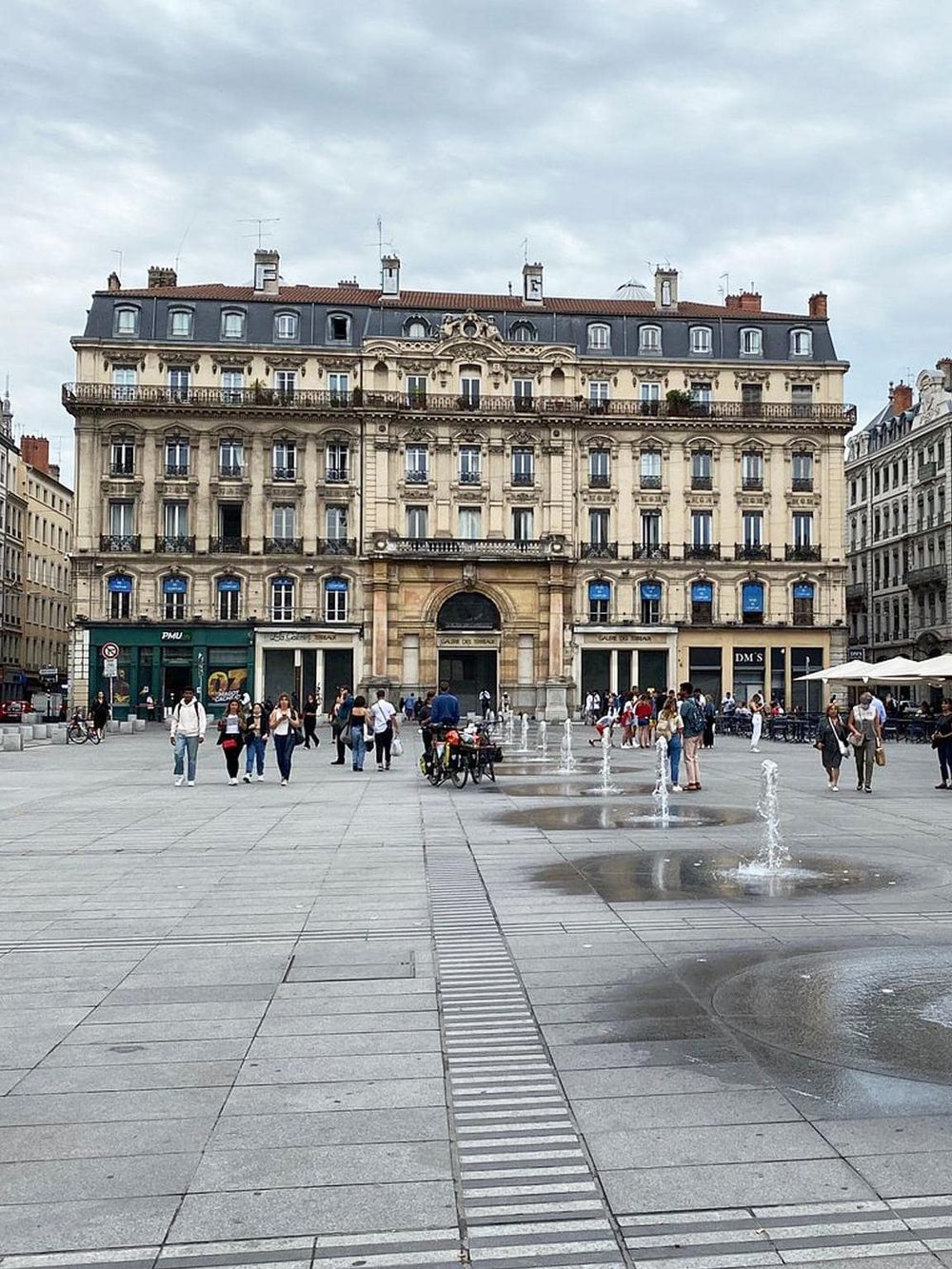 Historic Parisian building on bustling city square with fountains and pedestrians.