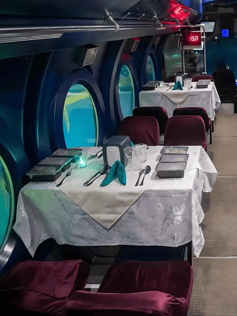 Elegant underwater-themed dining setup on a cruise ship with tableware, ambient lighting, and panoramic portholes.