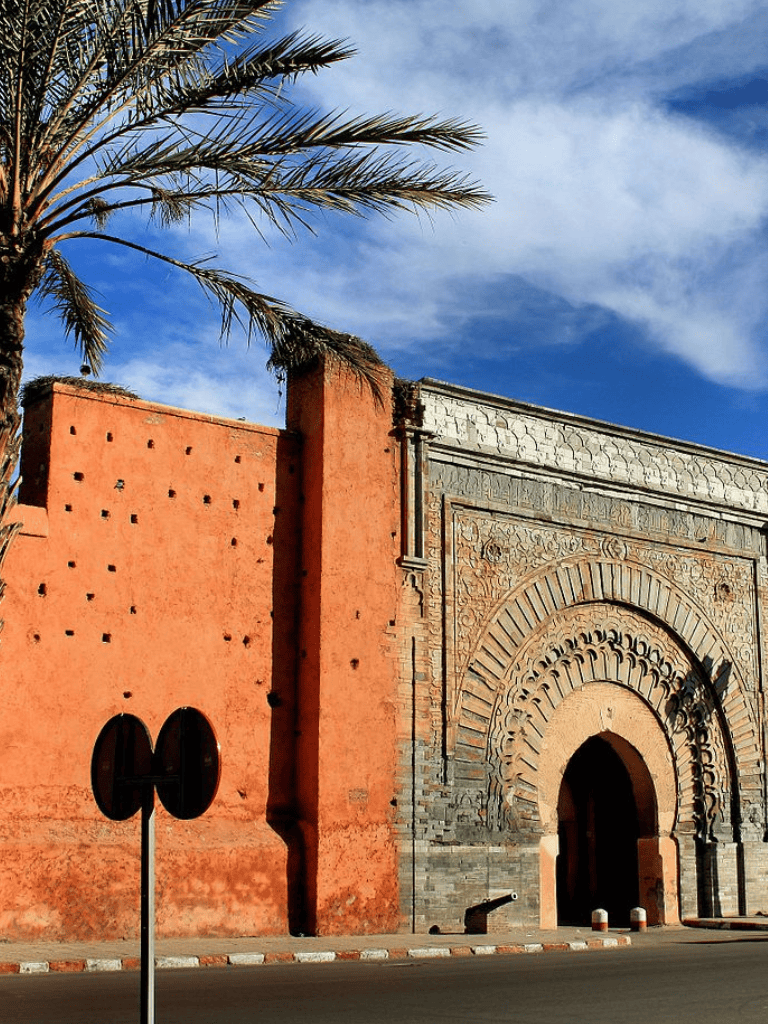 Vibrant Moroccan city gate with intricate architectural details and a palm tree against a blue sky.