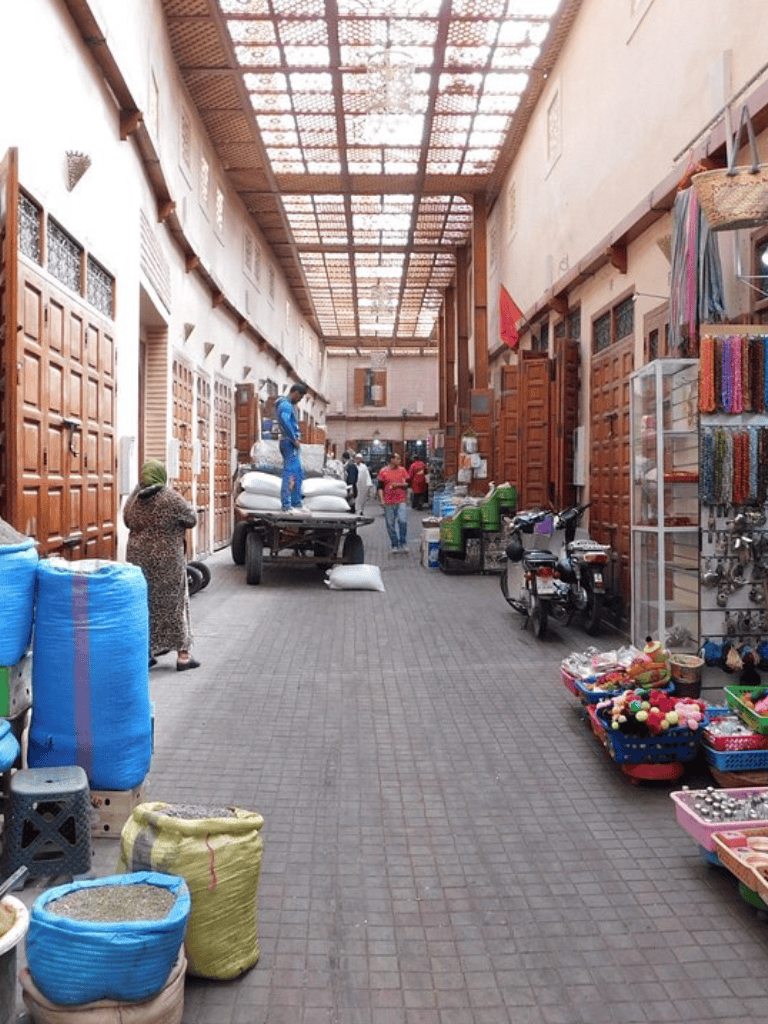 Colorful market street scene with goods for sale, including textiles, accessories, and grains, under a high wooden ceiling.