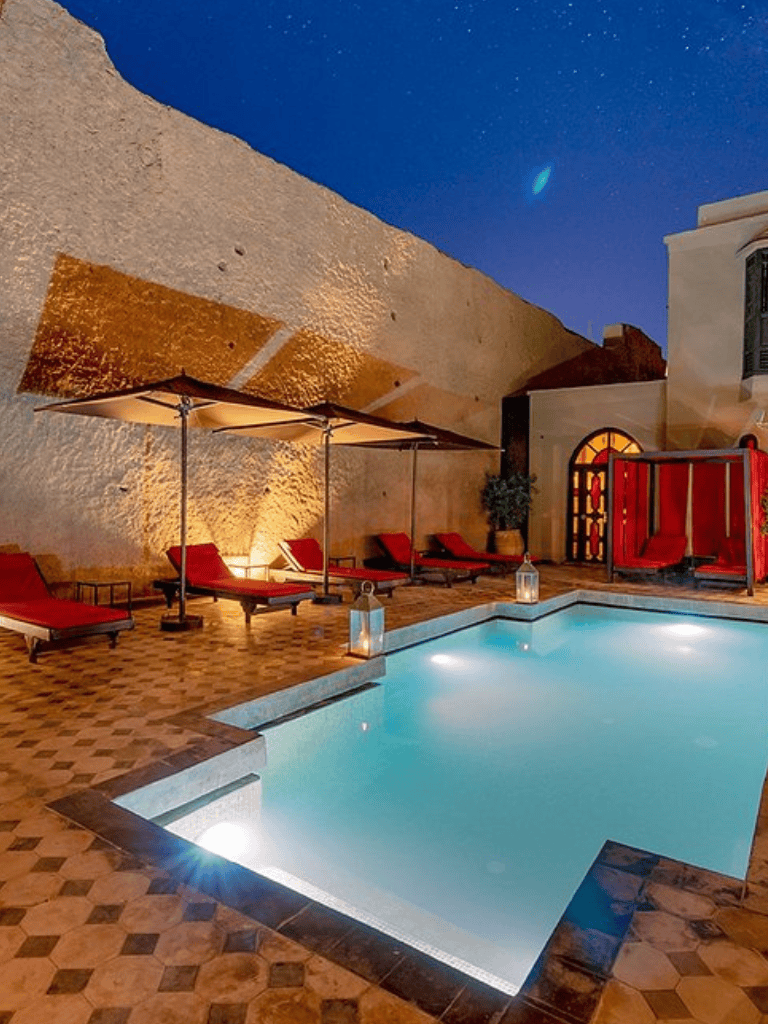 Luxurious outdoor pool at night with lounge chairs and umbrellas, cozy lighting, and historic architecture in the background.
