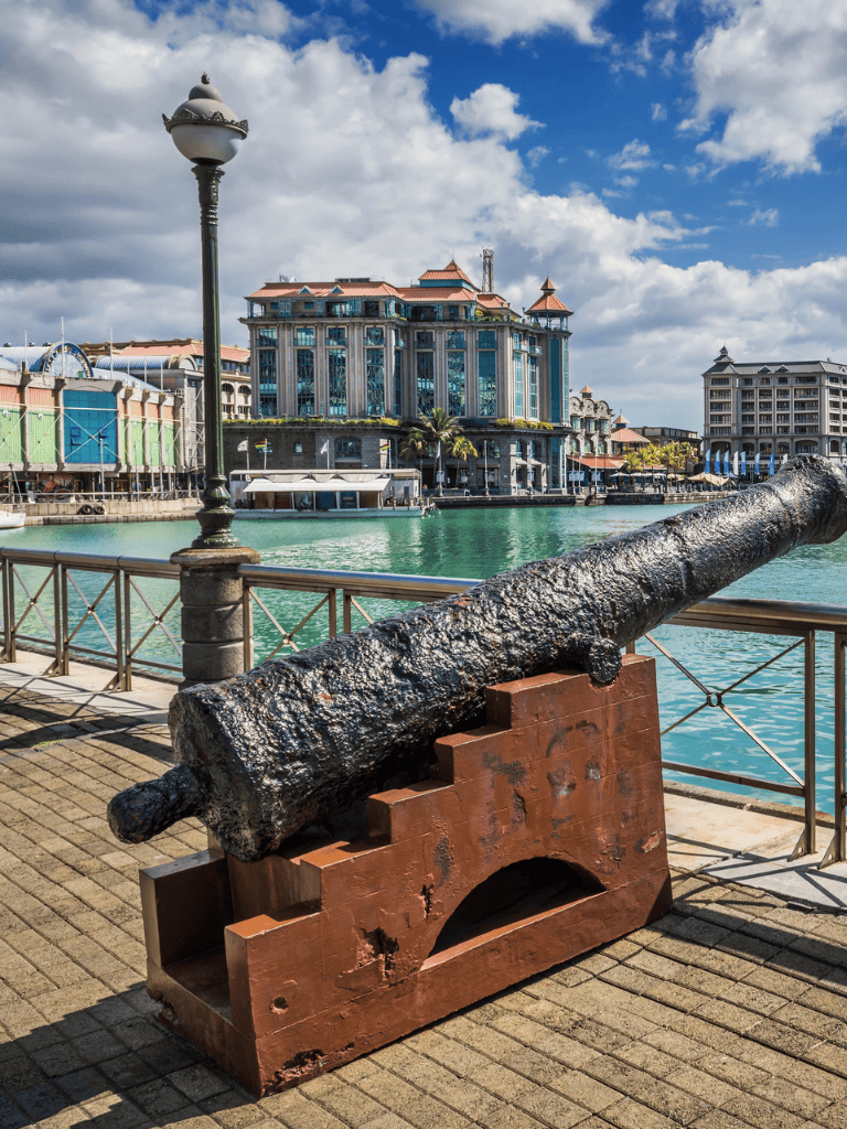 Old cannon on waterfront with colorful buildings and cloudy sky in the background, showcasing historic maritime artifacts.