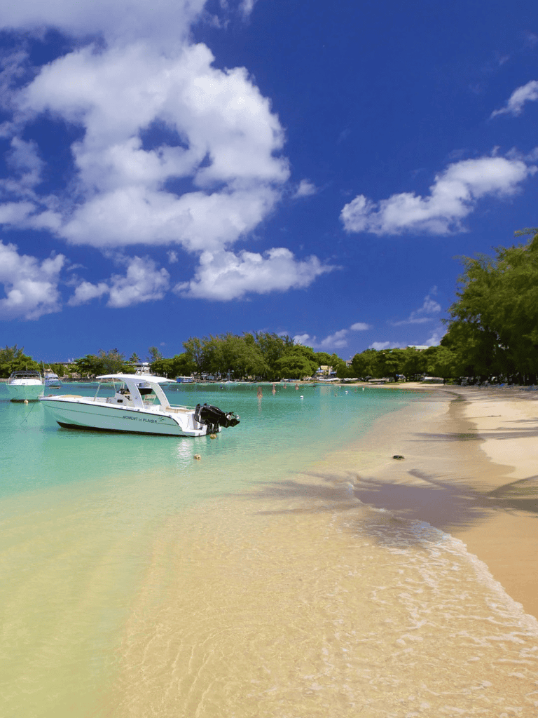 Serene beach with boats, clear turquoise water, and lush green trees under a blue sky with fluffy clouds.