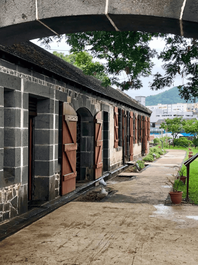 Aged stone building with wooden shutters, outdoor pathway, lush greenery, and modern cityscape in the background.
