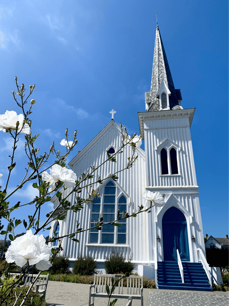 Traditional white wooden church with tall steeple under blue sky, spring flowers in foreground.
