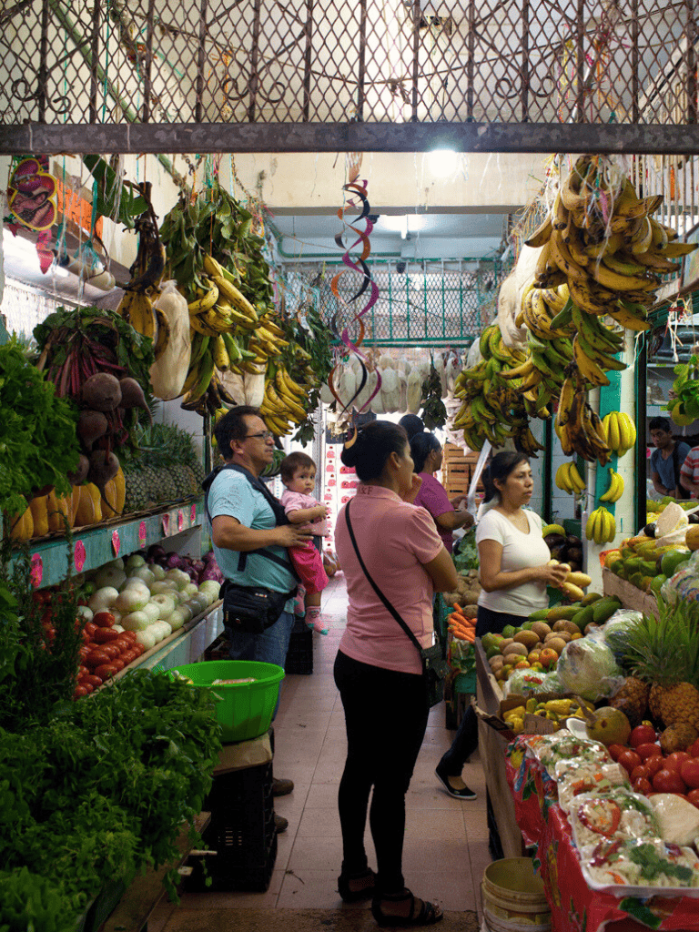 Colorful indoor marketplace with fresh fruits and vegetables, vibrant produce displays, diverse shoppers, and hanging bananas and produce.