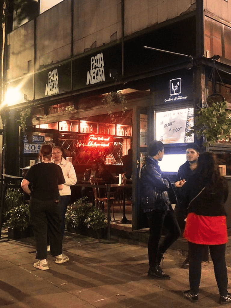 Nightlife restaurant exterior with people chatting, neon signs, and outdoor seating, representing a lively downtown dining scene.