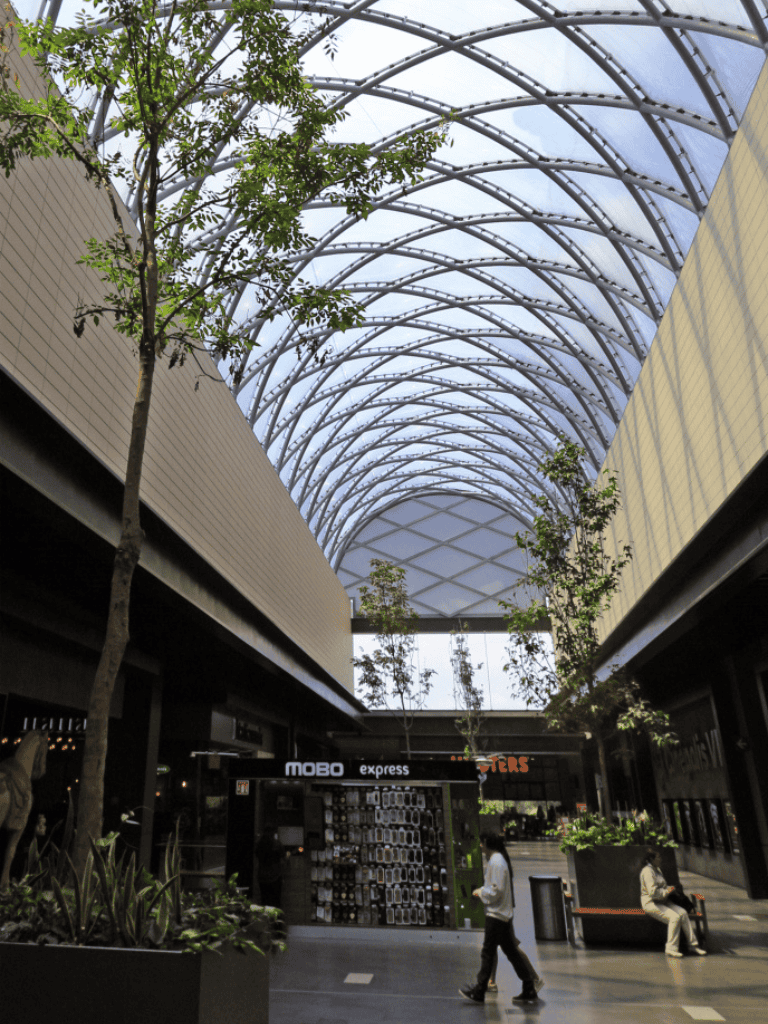 Indoor shopping mall with glass arch ceiling, retail stores, and greenery.