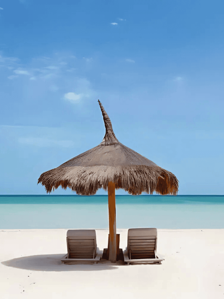 Tropical beach with straw umbrella and lounge chairs overlooking clear blue ocean.