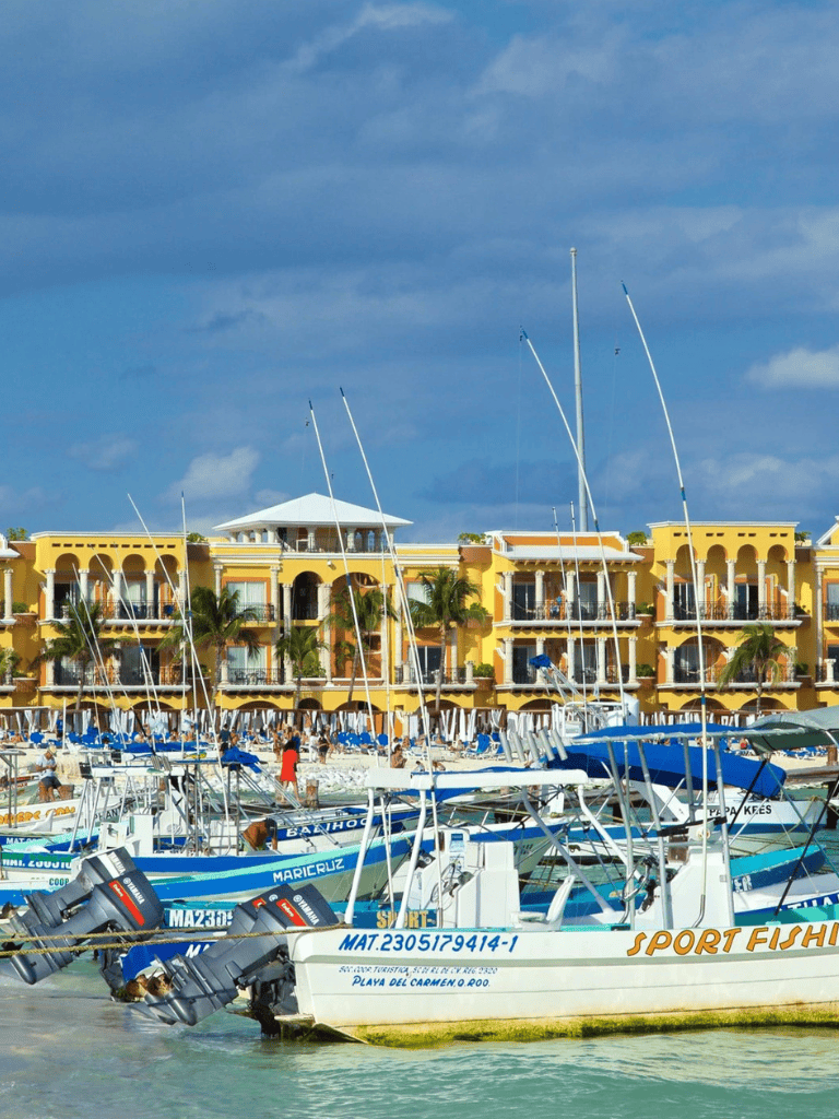Boats docked at marina near colorful resort with yellow buildings, sunny weather, and blue sky.