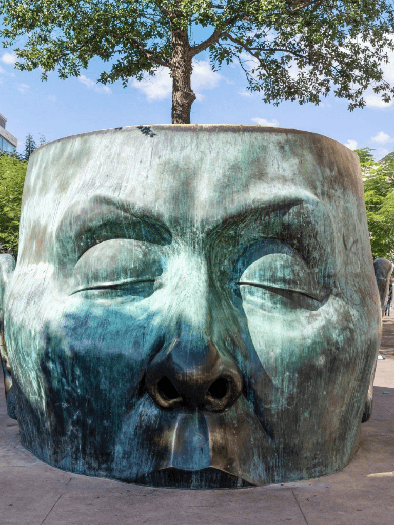 Ancient Buddha statue head sculpture in public park with trees and blue sky in the background.