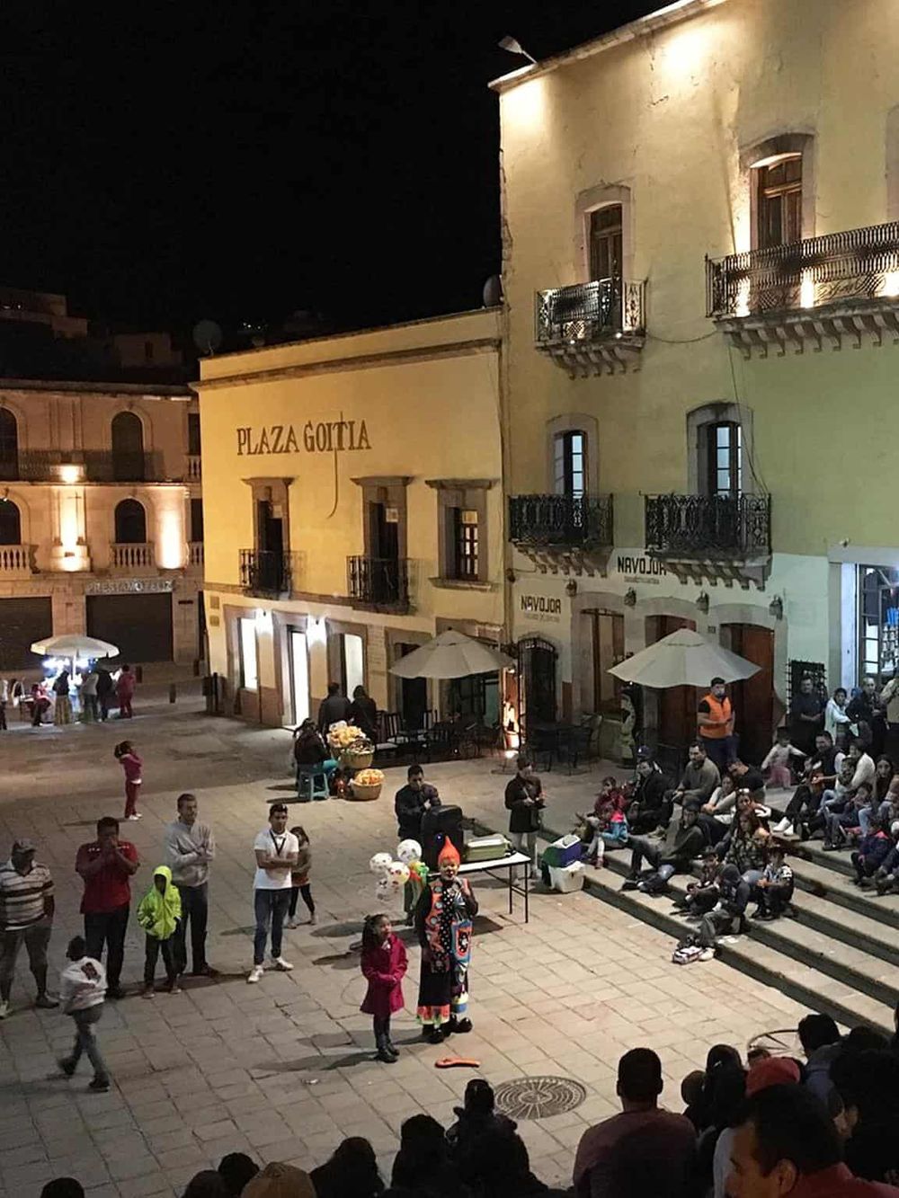 Nighttime scene at Plaza Gotta with street performers and audience in a historic town square.