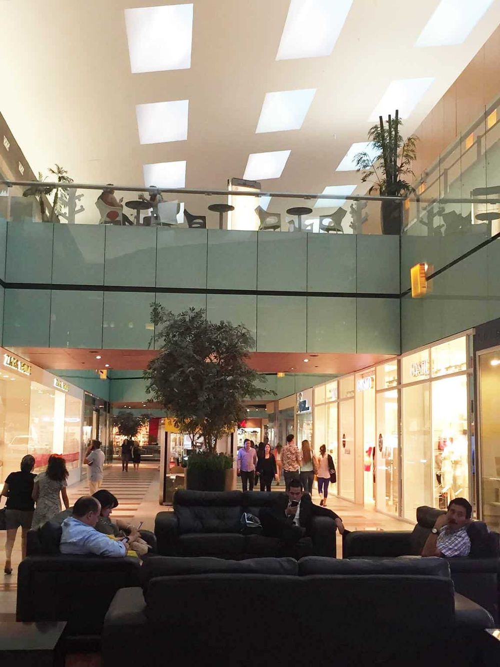 Bright modern shopping mall lobby with seating area, shops, and upper-level cafe, lively shopping and relaxation environment.