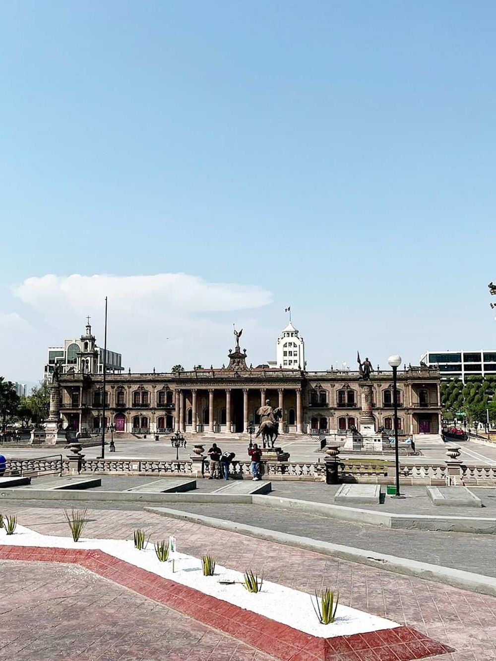 Historic city hall building with a statue in front, surrounded by urban scenery and clear blue sky.